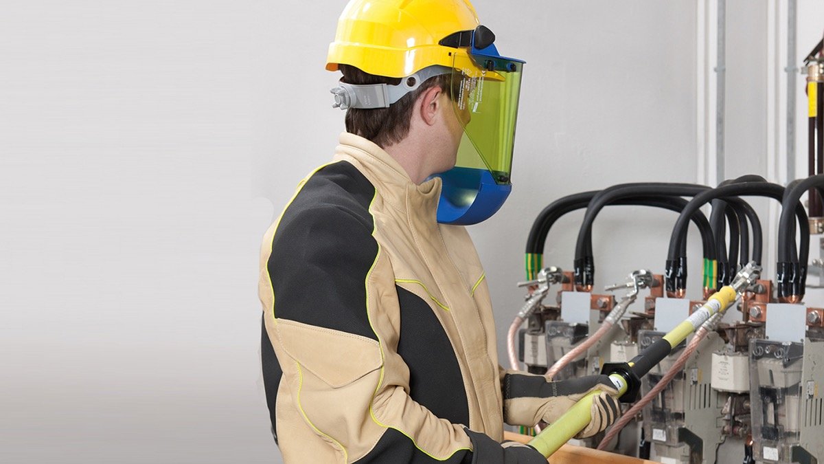 Person in Schutzkleidung, Helm und Visier arbeitet an einer elektrischen Schaltanlage mit isolierter Bedien- und Sicherheitsstange