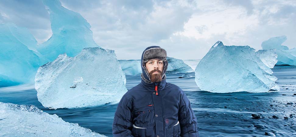 Mann trägt eine dunkelblaue Herren-Winterjacke und Fellmütze vor großen Eisblöcken in einer Gletscherlagune